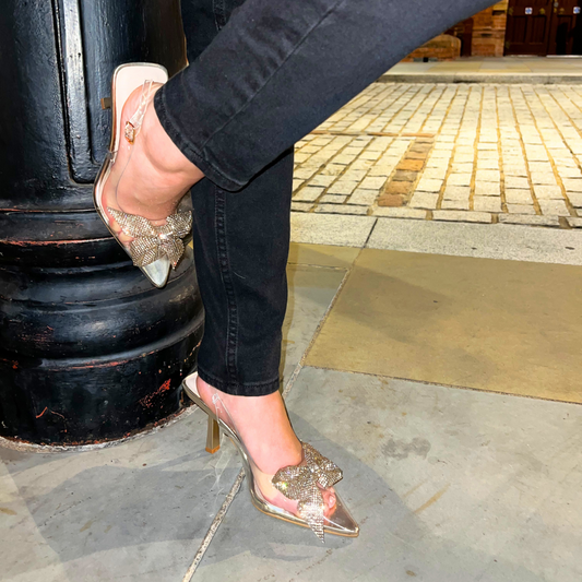 Woman's foot wearing stylish perspex court shoe with diamante brooch detail in gold, a comfortable and fashionable choice for London events.