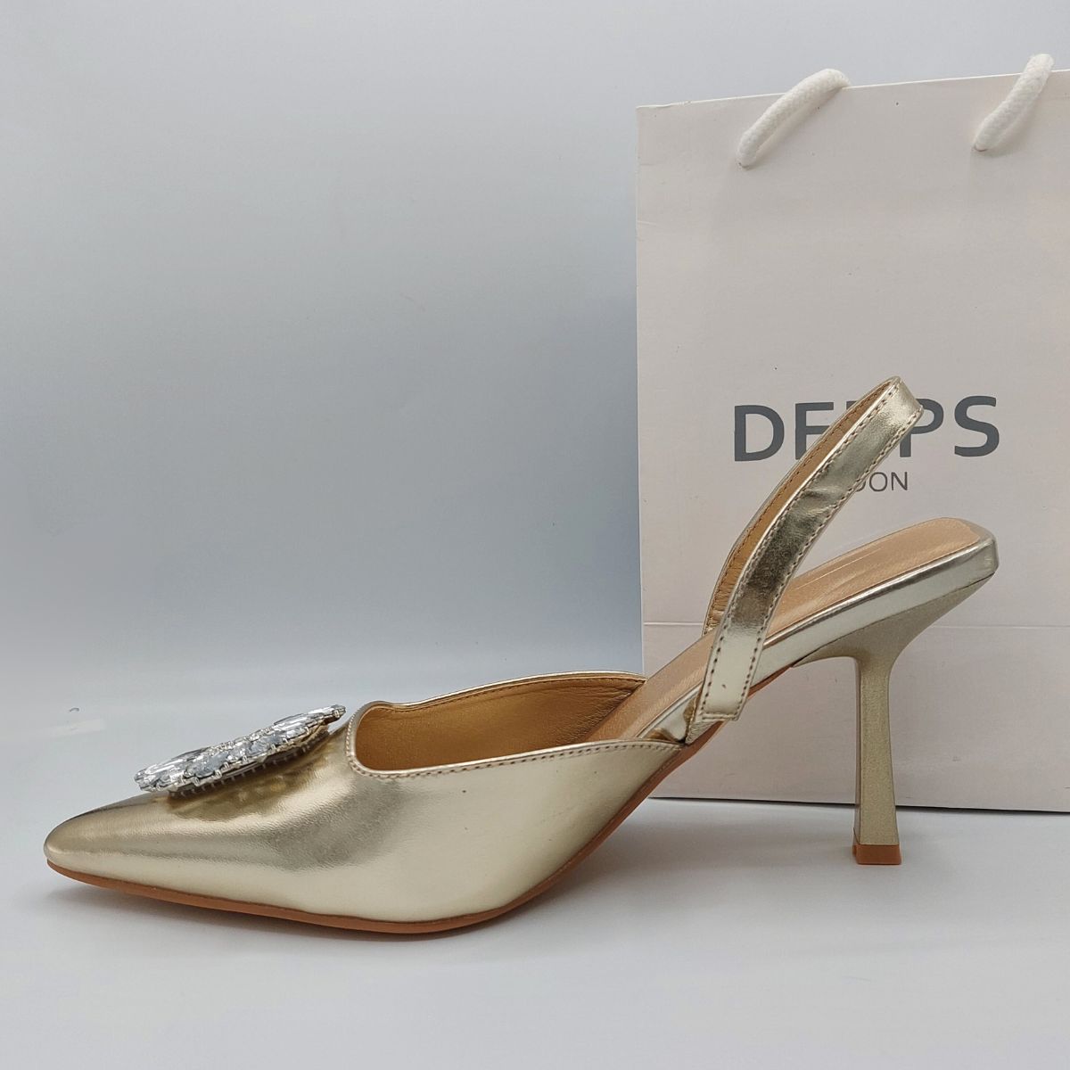 Woman's foot wearing stylish perspex court shoe with diamante brooch detail in Gold PU, a comfortable and fashionable choice for London events.