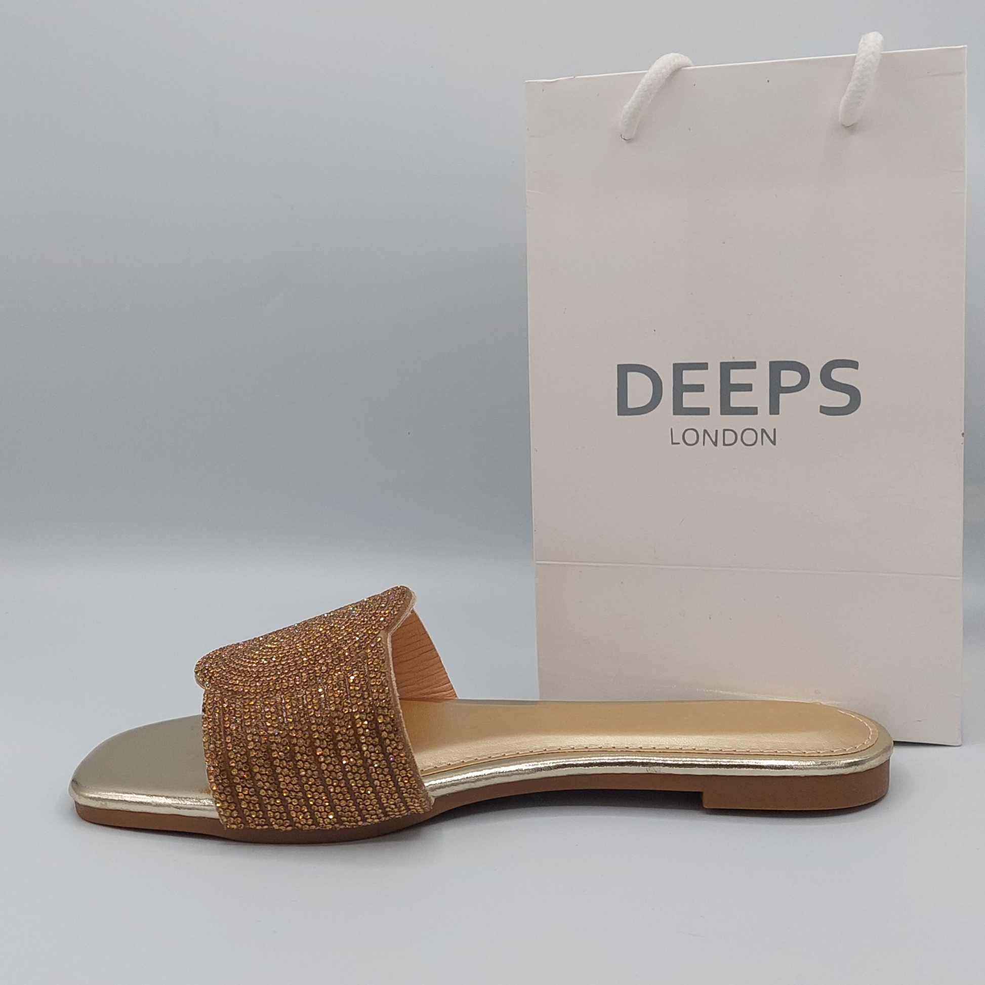 Woman's foot wearing stylish gold perspex flat slider sandals with diamante detail, a trendy London fashion statement.

