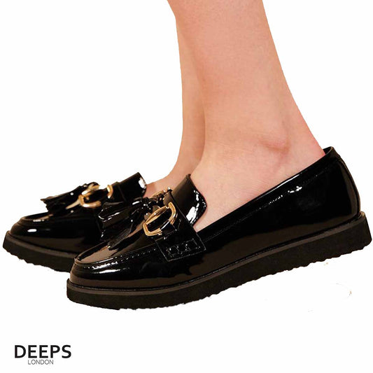 Woman's foot wearing stylish black patent classic slip-on loafers with a gold bar and tassels, a comfortable and fashionable choice for London life.